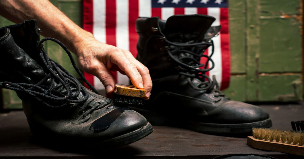 A hand brushing black shoe polish onto black military boots with an American flag in the background.
